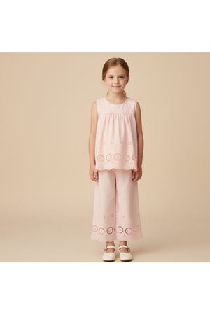 A young girl stands wearing a blush organic cotton eyelet top and matching pant set, featuring delicate embroidered details. The outfit is perfect for warm-weather outings and celebrations.