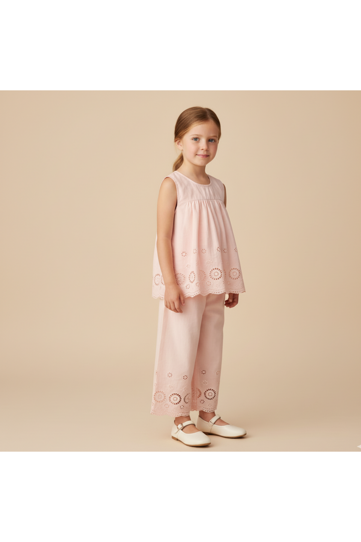 A young girl stands wearing a blush organic cotton eyelet top with delicate embroidered details paired with matching pants. The soft, breezy outfit is ideal for warm-weather occasions.