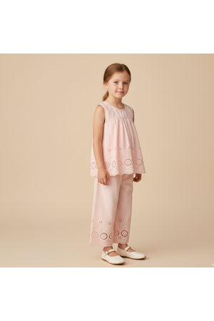 A young girl stands wearing a blush organic cotton eyelet top with delicate embroidered details paired with matching pants. The soft, breezy outfit is ideal for warm-weather occasions.