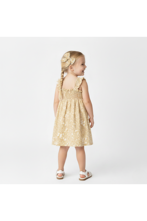 A young girl stands with her back to the camera, wearing a yellow floral smocked sundress. Her hair is styled in braids, adorned with a matching bow.