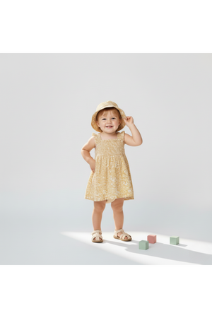A toddler wearing a yellow blossom smocked sundress stands on a light background, smiling and wearing a matching sun hat. Colorful blocks are scattered nearby, enhancing the playful scene.