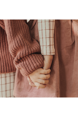 Two children hold hands, showcasing a cozy knitted mauve sweater with a textured pattern and a patterned shirt featuring a light check design. The soft fabrics emphasize warmth and togetherness.