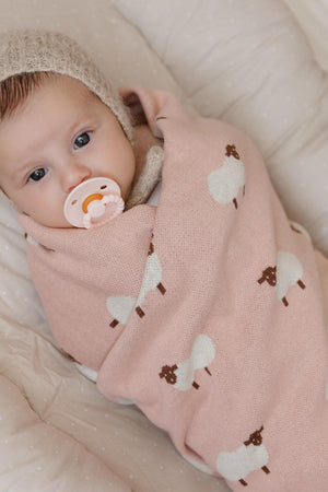 A baby wrapped in a pink swaddle with sheep patterns, wearing a knitted hat and holding a pacifier.
