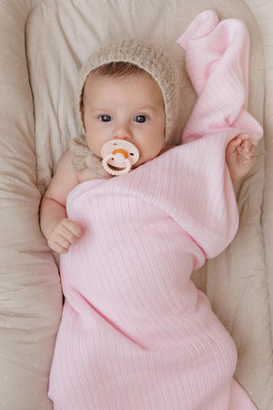 A baby wrapped in a pink ribbed knit blanket, wearing a beige knit hat and holding a pacifier.