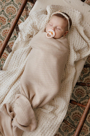 A baby sleeps peacefully in a beige ribbed knit swaddle blanket, lying on a textured cream blanket.