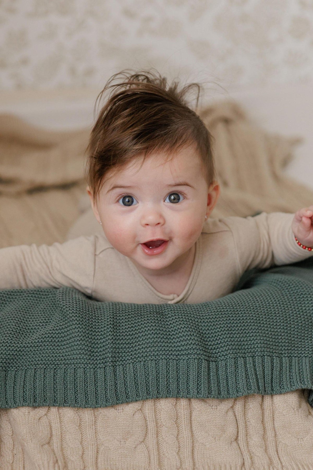 A baby with dark hair and blue eyes on a green knit blanket, wearing a beige outfit.