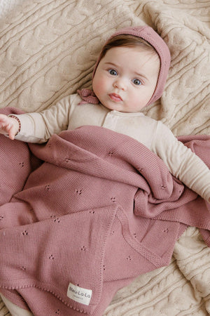 A baby wearing a beige outfit and pink bonnet is wrapped in a pink knit blanket on a textured beige.