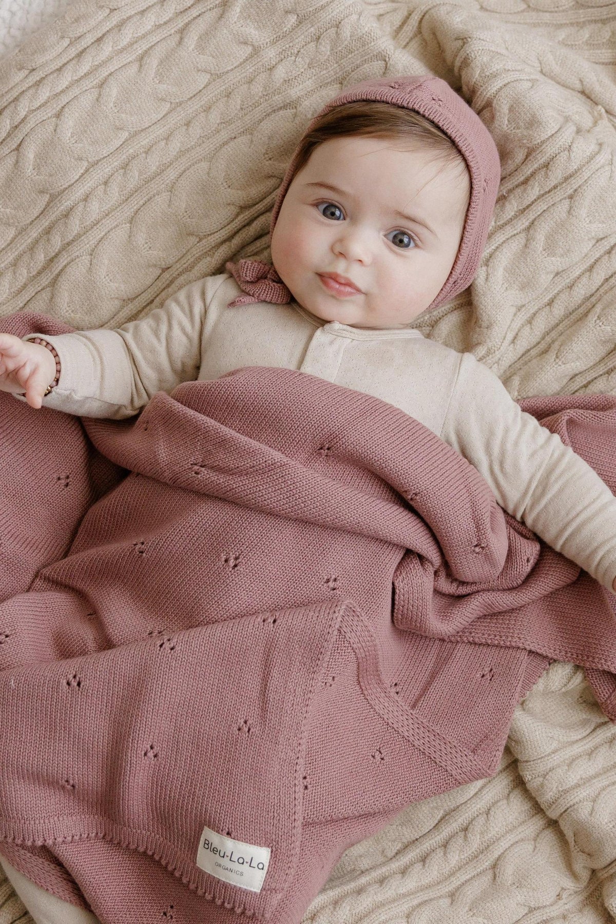 A baby wrapped in a pink organic pointelle blanket with a matching hat, lying on a beige textured surface.