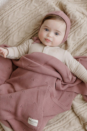 A baby wrapped in a pink organic pointelle blanket with a matching hat, lying on a beige textured surface.