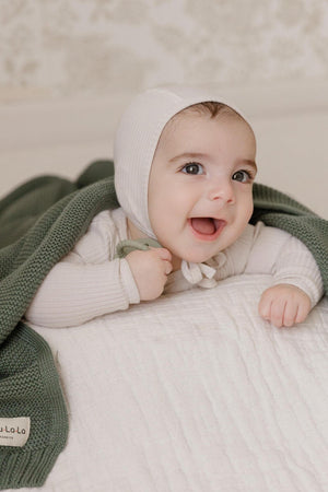 A smiling baby wearing a white bonnet and lying on a green knit blanket.