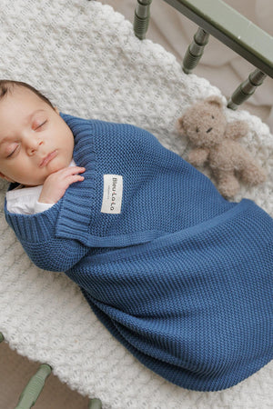 A baby sleeps in a blue knit swaddle with a label, lying on a textured white blanket with a teddy.