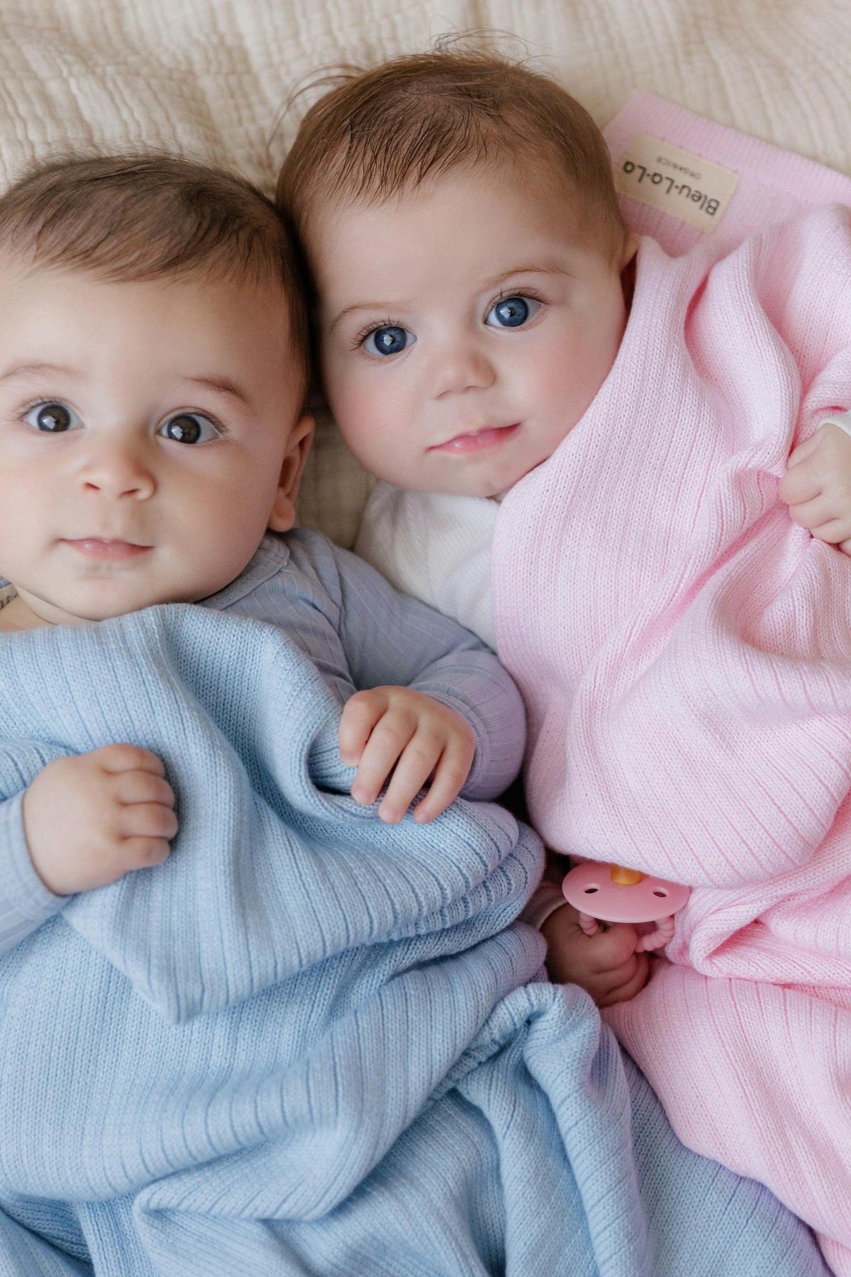 Two babies wrapped in ribbed knit blankets, one blue and one pink, lying on a soft surface.