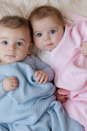 Two babies wrapped in ribbed knit blankets, one blue and one pink, lying on a soft surface.