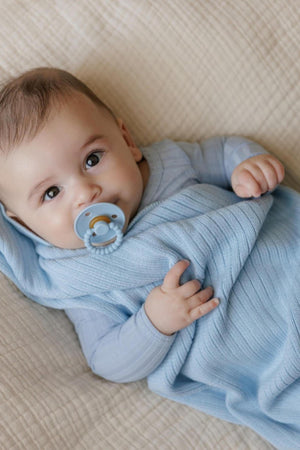 A baby wrapped in a soft, blue ribbed knit swaddle, holding a pacifier, lies on a beige blanket.