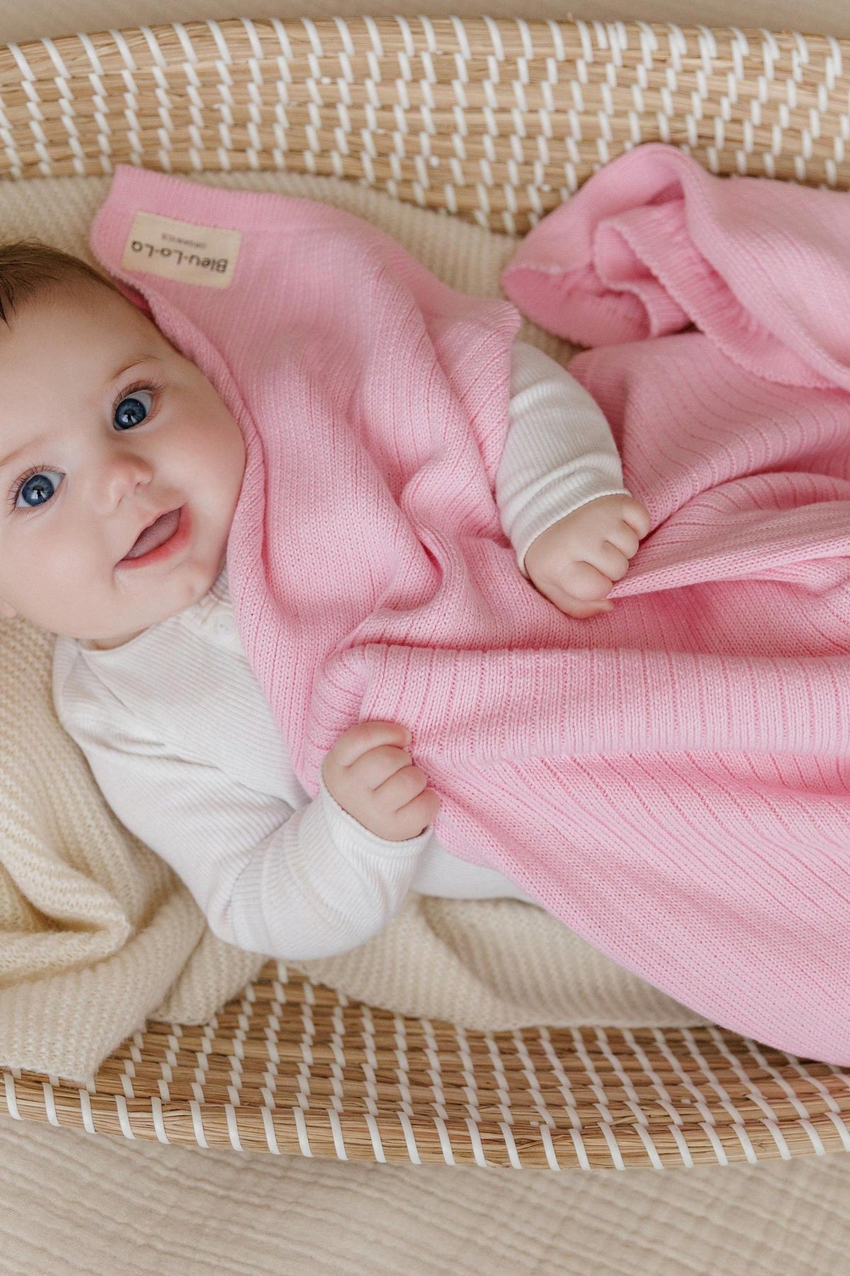 A baby lies in a woven basket, wrapped in a pink ribbed knit blanket, smiling warmly.