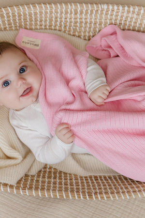 A baby lies in a woven basket, wrapped in a pink ribbed knit blanket, smiling warmly.