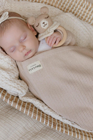 A baby sleeps in a beige ribbed knit swaddle, holding a knitted bear toy, lying in a woven basket.