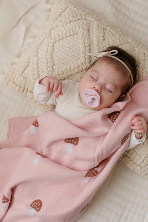 A baby sleeps peacefully with a pink mushroom-patterned swaddle blanket, lying on a textured cream pillow.