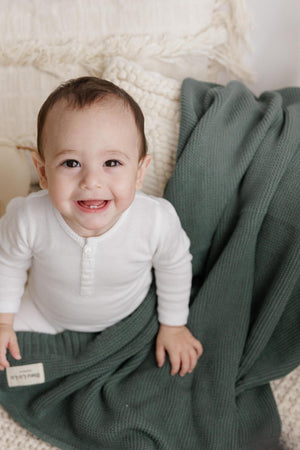 A smiling baby in a white outfit sits on a soft, green knit blanket, with textured cushions behind.