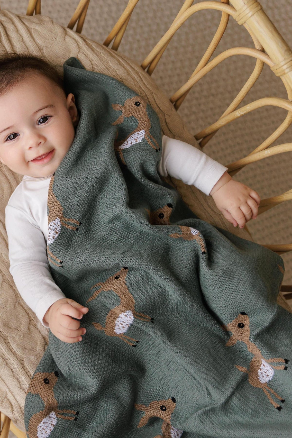 A baby wrapped in a green knit blanket with deer patterns, lying on a woven chair, smiling.
