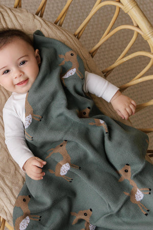 A baby wrapped in a green knit blanket with deer patterns, lying on a woven chair, smiling.