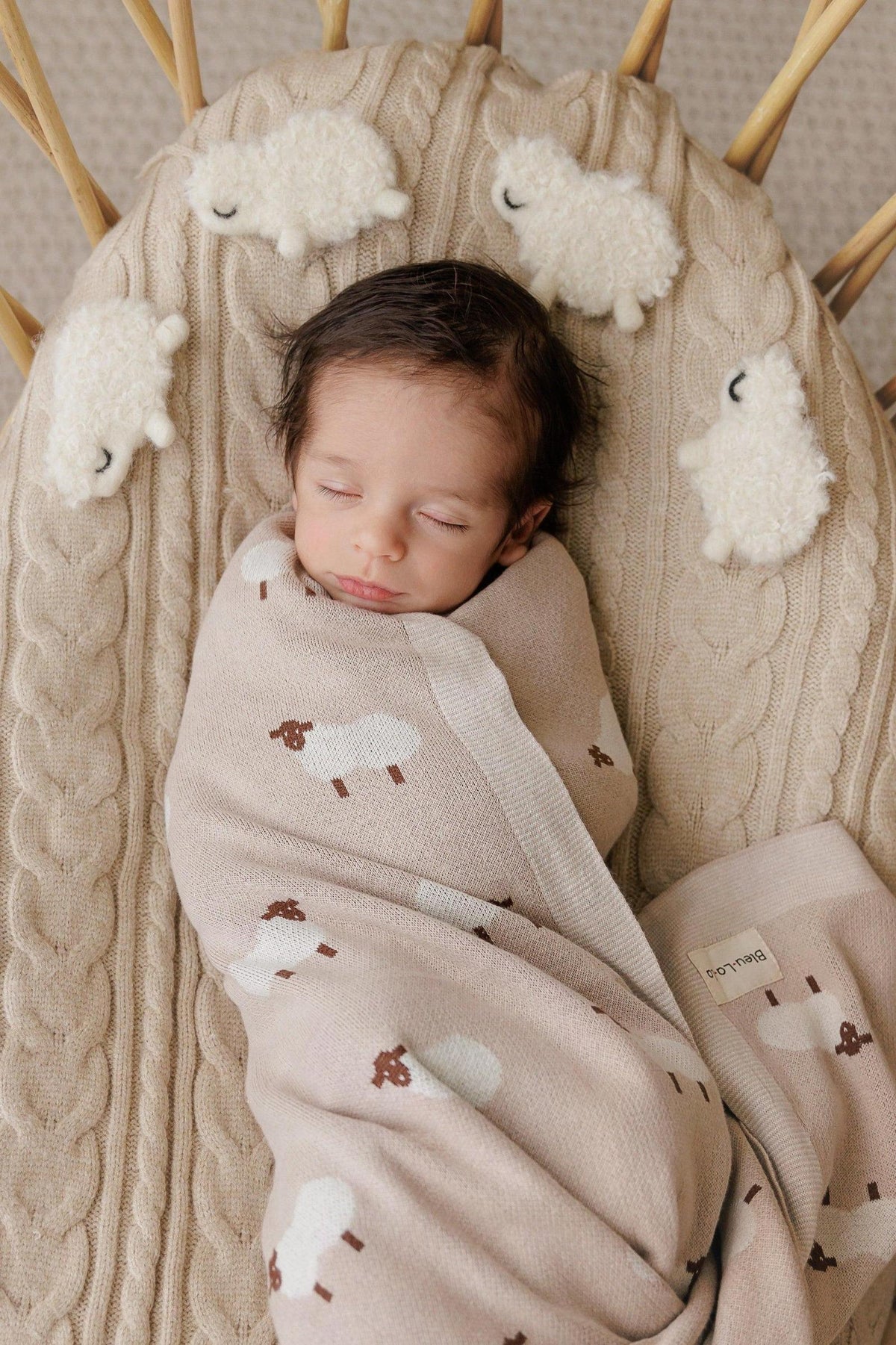 A baby sleeps peacefully, swaddled in a beige blanket with sheep patterns, on a textured cream cushion.