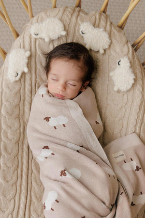 A baby sleeps peacefully, swaddled in a beige blanket with sheep patterns, on a textured cream cushion.