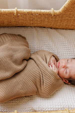 A baby wrapped in a brown knit swaddle blanket rests peacefully in a woven basket.