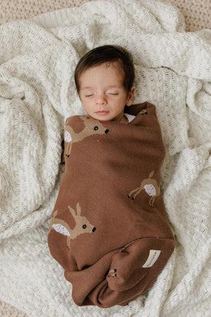 A baby wrapped in a brown swaddle with deer patterns, lying on a textured white blanket.
