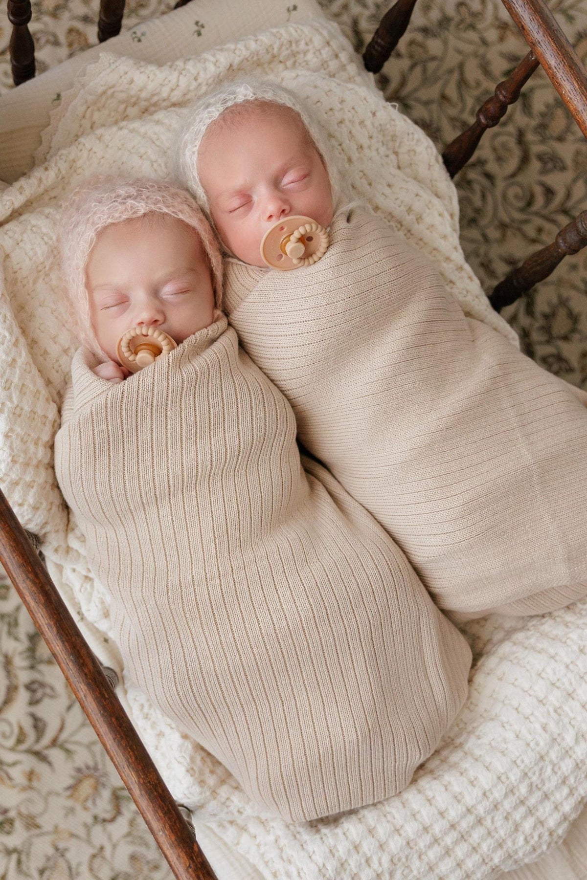 Two babies peacefully sleeping in ribbed knit swaddle blankets on a textured cream blanket.