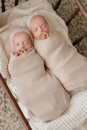 Two babies peacefully sleeping in ribbed knit swaddle blankets on a textured cream blanket.