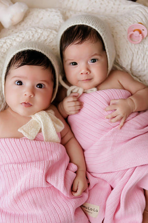 Two babies in pink ribbed knit blankets and cream hats, lying on a soft, textured surface.