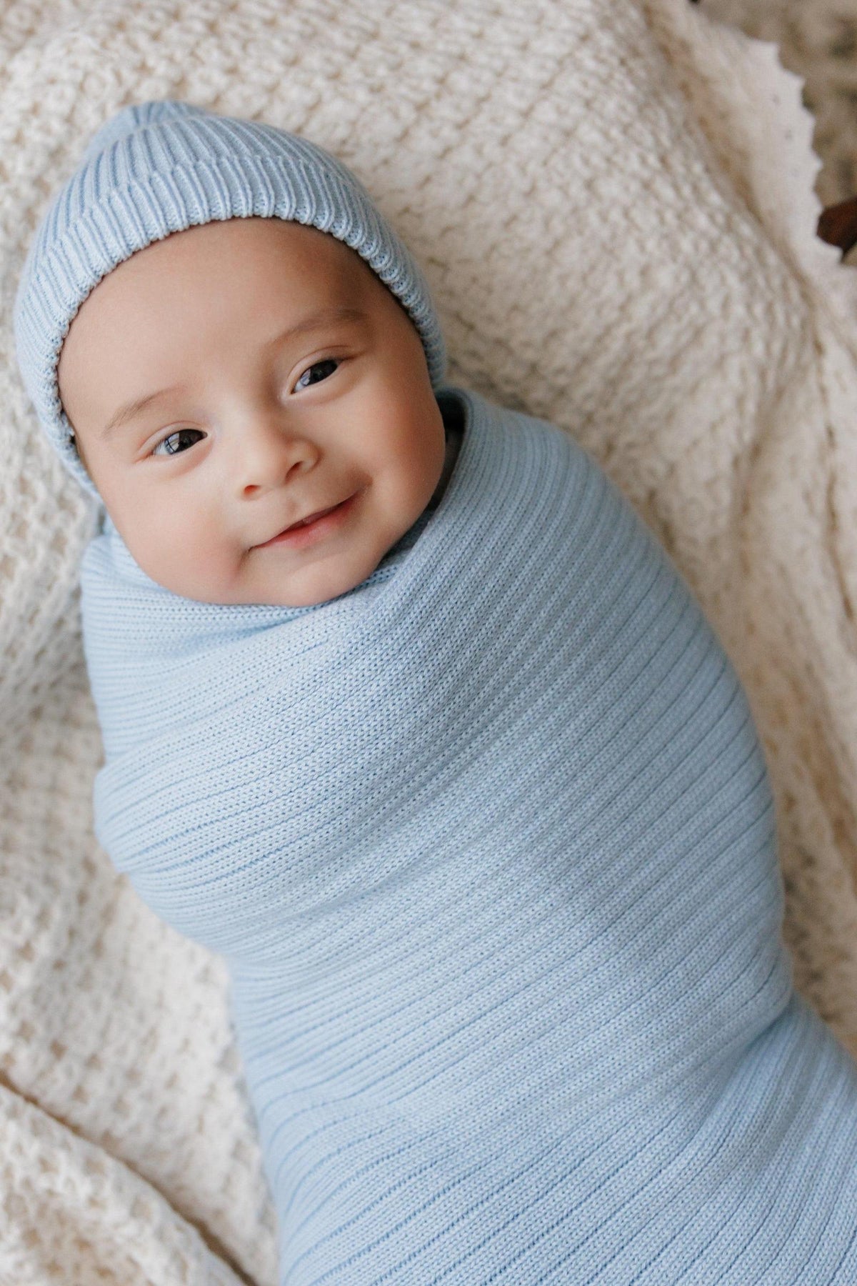 A baby wrapped in a light blue ribbed knit swaddle blanket with a matching hat, lying on a textured surface.