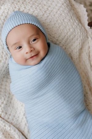 A baby wrapped in a light blue ribbed knit swaddle blanket with a matching hat, lying on a textured surface.