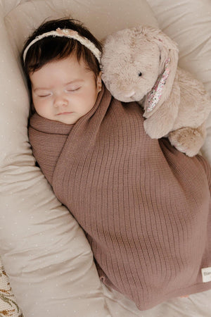 A baby sleeps wrapped in a brown ribbed knit blanket with a plush bunny, lying on a soft cushion.