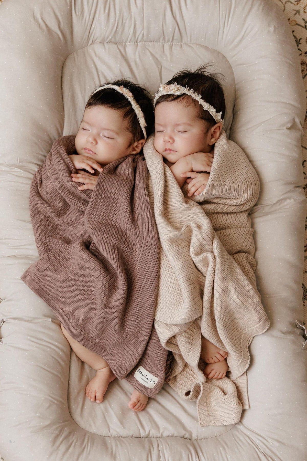 Two sleeping babies in ribbed knit swaddle blankets, one brown and one beige, lying on a soft cushion.