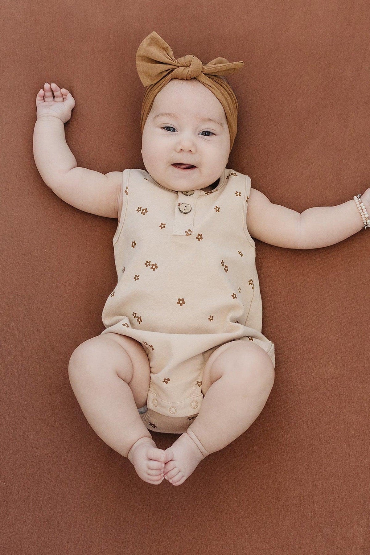 A baby lies on a rust-colored crib sheet, wearing a beige floral romper and a matching headband.