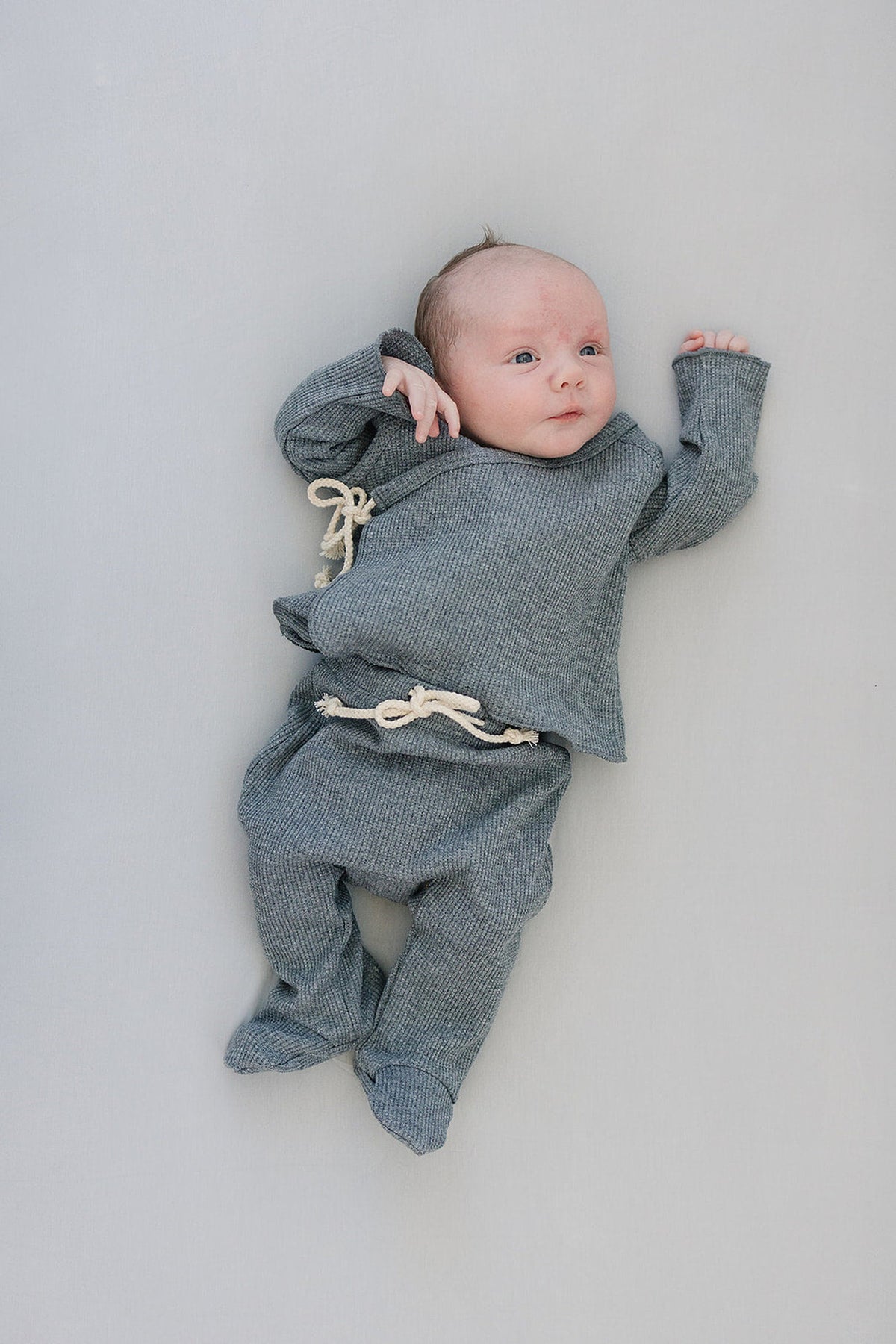 A baby lies on a light surface wearing a gray knit outfit with drawstring details, looking content.