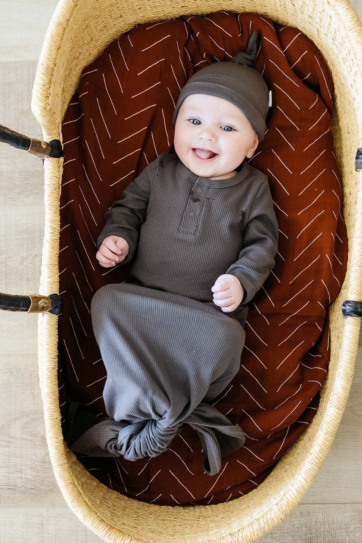 A baby in a charcoal ribbed knot gown and matching hat lies in a wicker basket with a brown cushion.