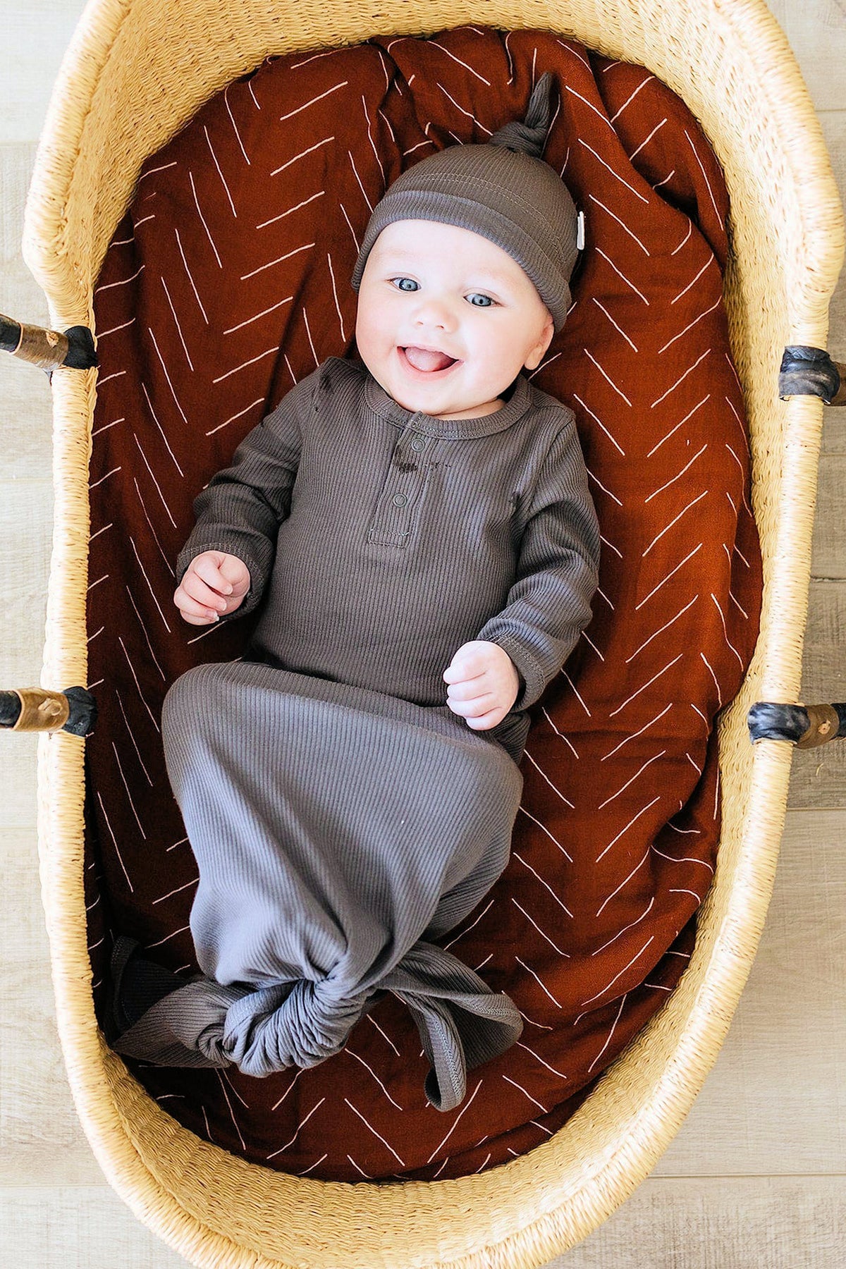 Smiling baby in a charcoal ribbed gown and knot hat, lying in a wicker basket with patterned cushion.