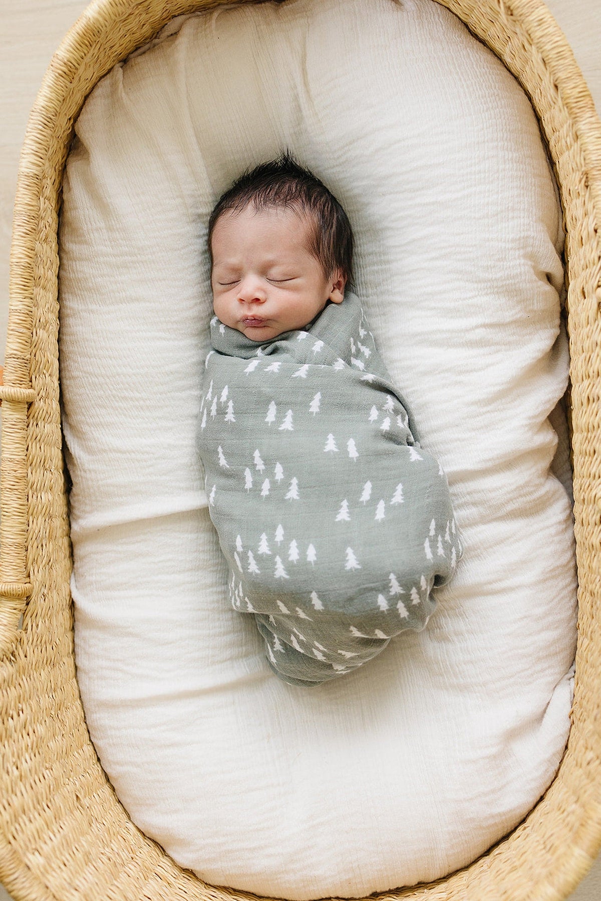 Newborn swaddled in a green muslin blanket with white tree patterns, lying in a woven basket.
