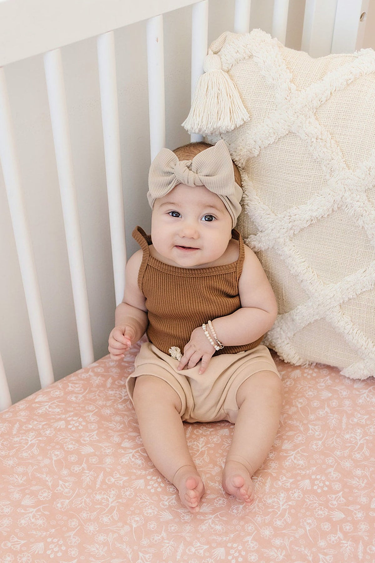 A baby wearing a beige head wrap and brown outfit sits in a crib with a pink floral sheet.