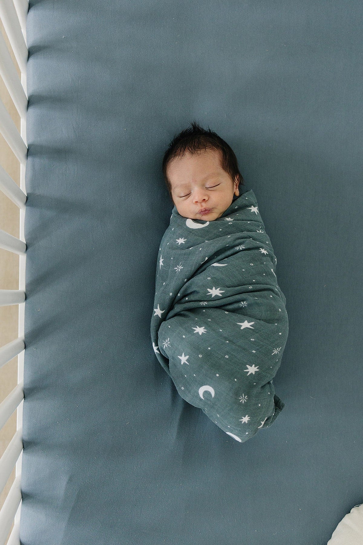 A newborn baby is swaddled in a star-patterned blanket, lying on a dusty blue crib sheet.