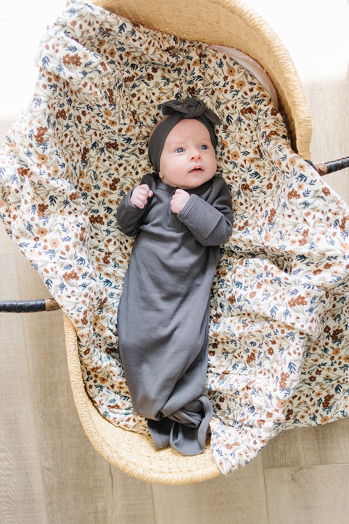 Newborn in a charcoal ribbed gown and matching headband, lying in a floral-lined basket on a wooden floor.