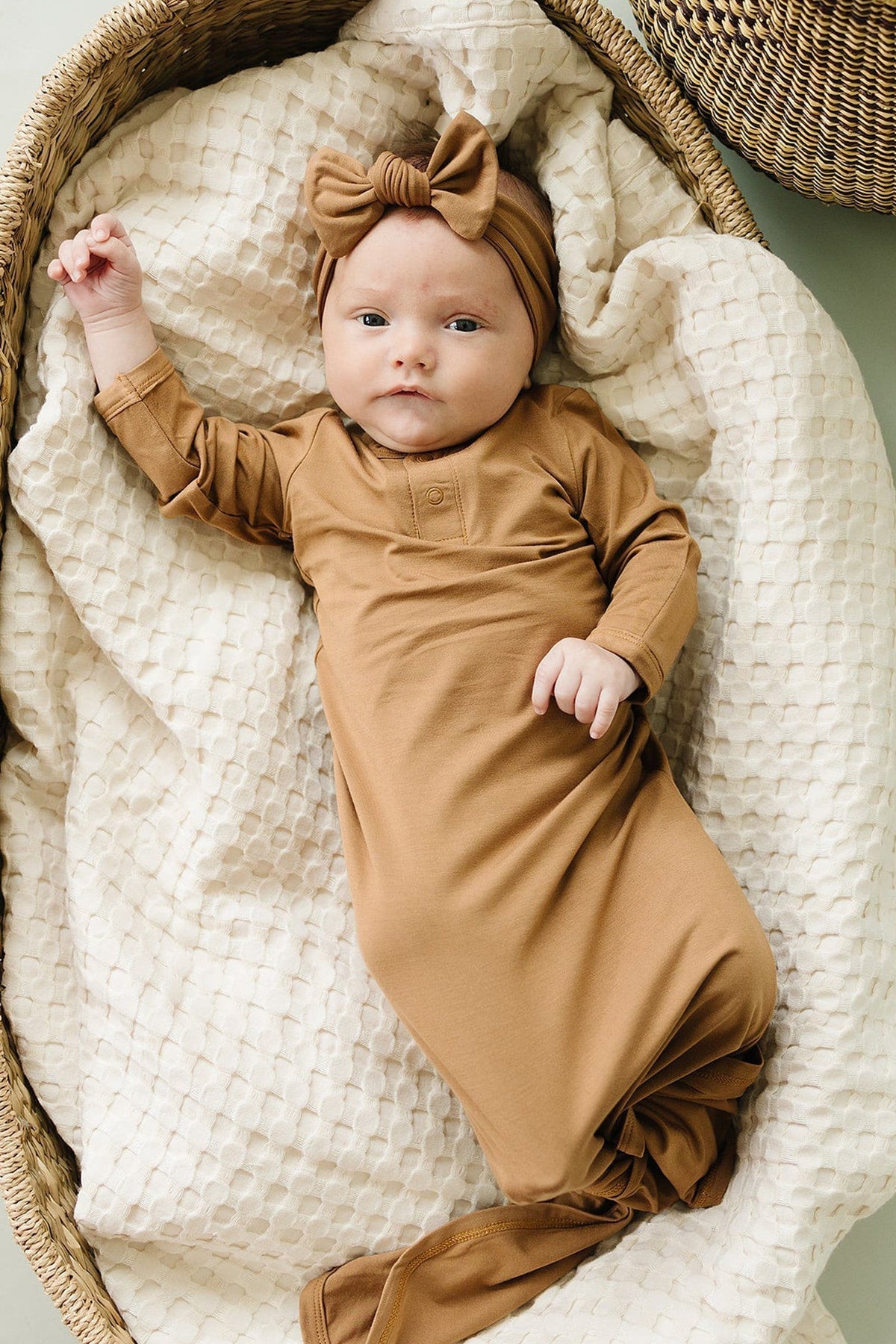 A baby in a mustard-colored bamboo gown with a matching headband lies on a textured white blanket.