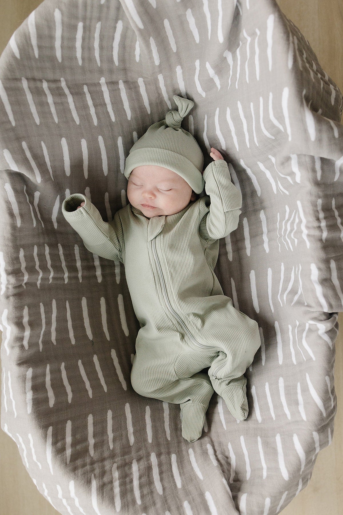 A baby wearing sage green ribbed pajamas and a matching hat lies on a patterned gray blanket.
