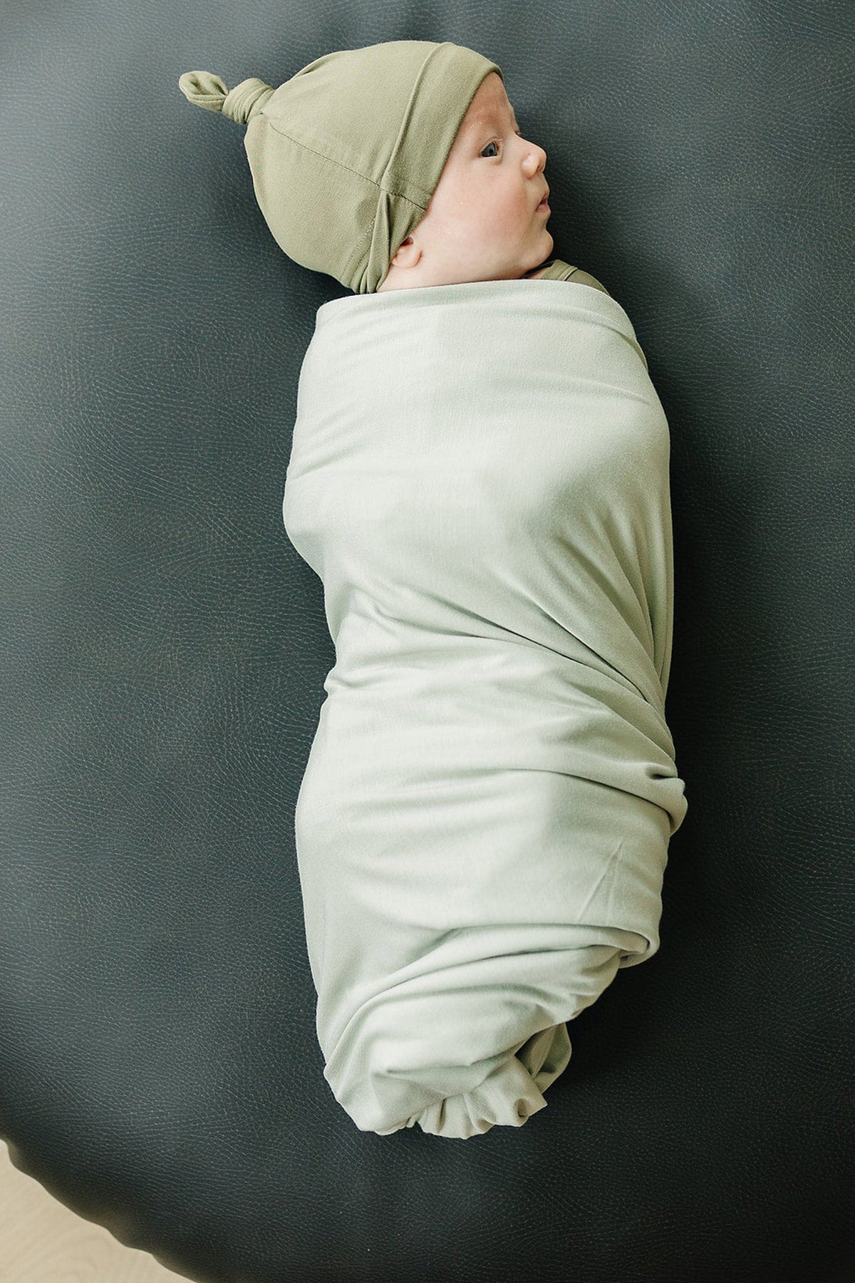 A baby wrapped in a light sage swaddle, wearing a matching knotted hat, lies on a dark surface.