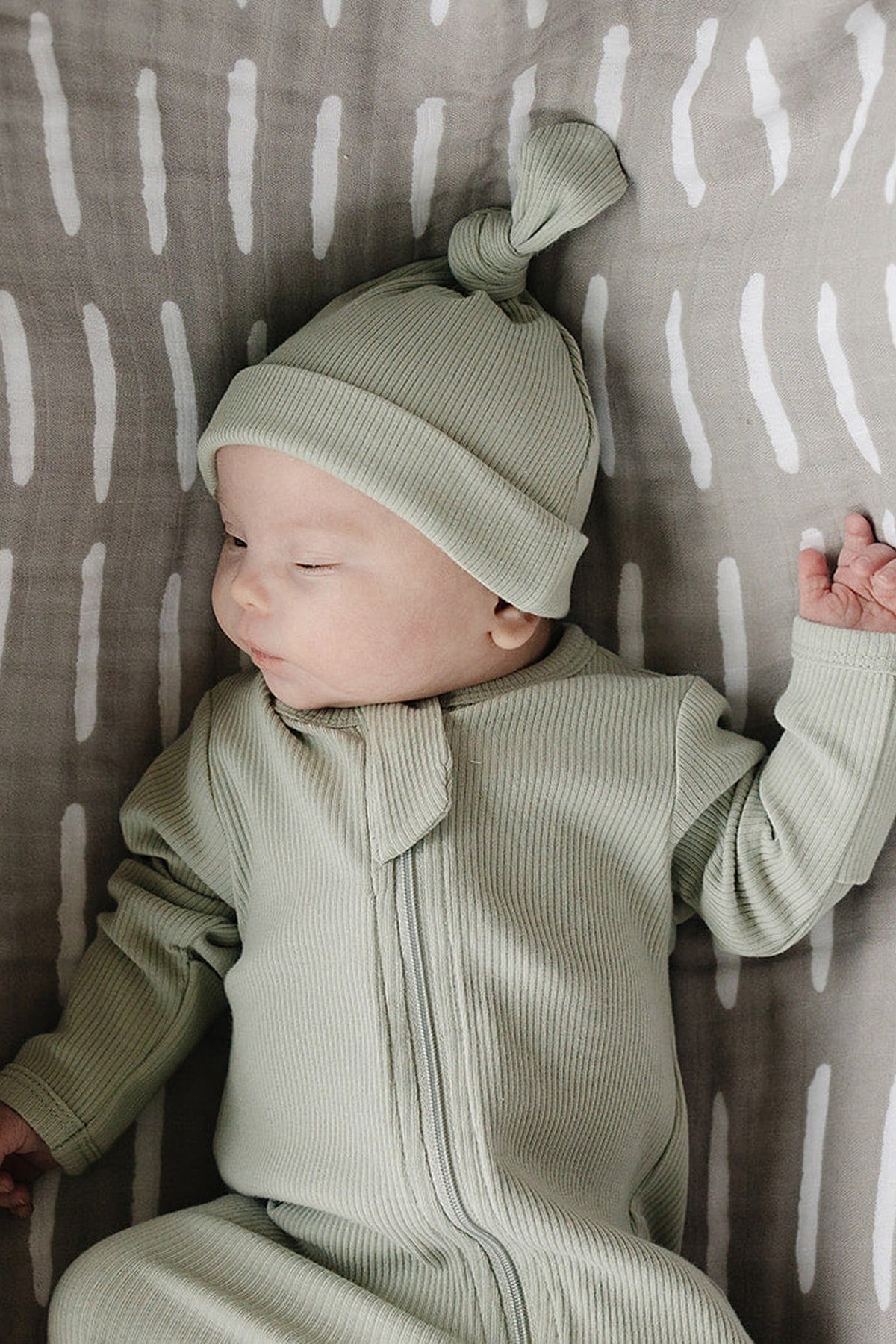 A baby wearing a sage ribbed knot hat and matching onesie lies on a patterned blanket.