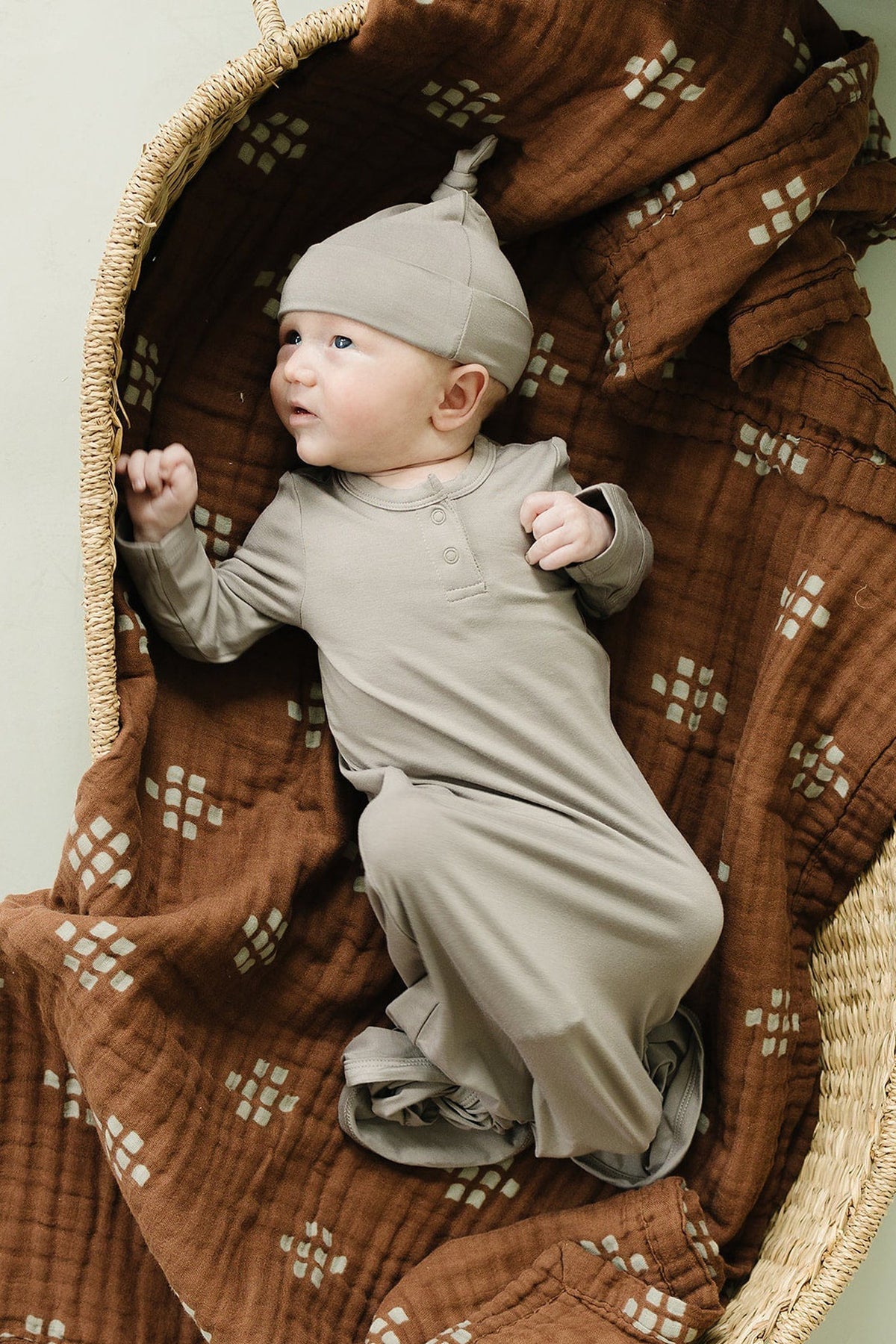 A baby in a beige swaddle and hat lies in a wicker basket on a patterned brown blanket.