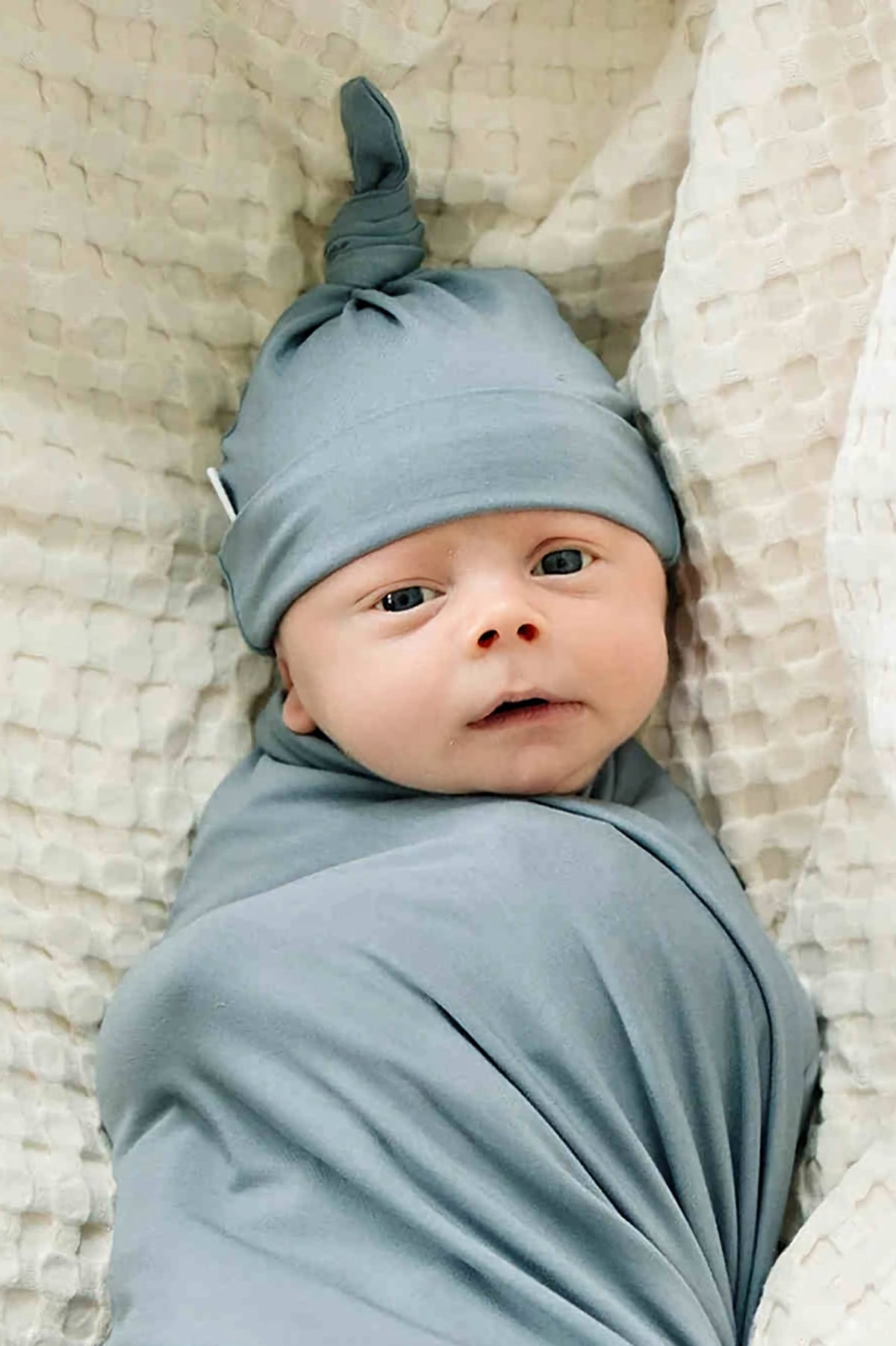 Newborn wearing a dusty blue knot hat and swaddle, lying on a textured cream blanket, looking upward.